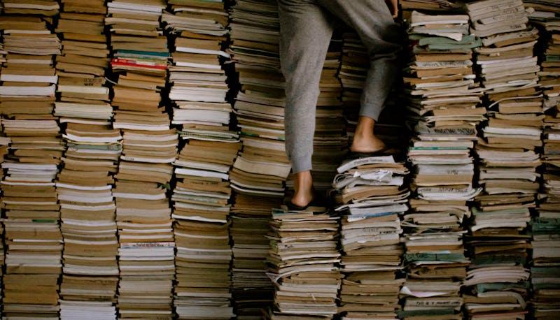 Uno studente si arrampica su una pila gigantesca di libri in cerca di un'idea per il Capolavoro